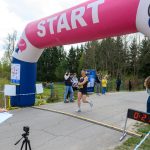 Biegacz przekracza linię mety pod różowo-niebieskim łukiem z napisem START. Obok stoi zegar pokazujący 21:22. W tle widoczne drzewa i kilka osób.