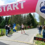 Na zdjęciu widać biegacza przekraczającego start pod różowym balonem z napisem. Tło stanowi zielony las i niebo. Po bokach stoją kibice obserwujący wyścig.