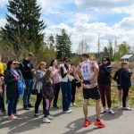 Na zdjęciu widać biegacza w kolorowej koszulce otoczonego przez ludzi. W tle są drzewa i niebo z chmurami. Grupa osób stoi na asfalcie, obserwując, co się dzieje.