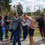 Mężczyzna zakłada medal biegaczowi w kolorowej koszulce. Wokół stoją osoby, które patrzą i robią zdjęcia. Tło to zalesiony teren z widocznym niebem.