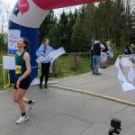 Biegaczka przekracza linię mety, nosząc zieloną koszulkę i numer 74. Za nią łuk z napisem Ale czas!, a tło stanowi zieleń drzew. Dwie osoby trzymają taśmę mety, a obok stoi kilka osób.