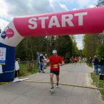 Zawodnik w czerwonej koszulce przekracza linię startu/metry. Nad nim różowy łuk z napisem START. W tle widać biegaczy i drzewa.