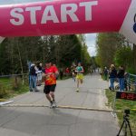 Biegacz przekracza linię mety pod różowo-białym łukiem z napisem START. W tle widać drzewa i innych biegaczy. Obok stoi osoba z zegarem o wyniku 25:17.