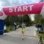 Biegaczka w zielonych spodenkach przekracza linię startu pod różowym namiotem. W tle zielone drzewa i kibice. Po prawej cyfrowy zegar z czasem.