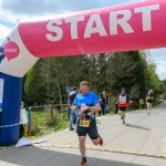 Biegacze przekraczają linię startu mety z różowym napisem START. W tle drzewa i kibice przy drodze. Dzień słoneczny, niebo chmurne.