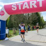 Biegacze na starcie pod różowym łukiem z napisem START. Widoczni są kibice na tle zieleni. Obok łuku stoi tablica z mapą.