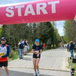 Biegacze przekraczają różowy łuk startowy na wiejskiej drodze, otoczeni drzewami. Tablica czasowa wskazuje 25 minut. Na poboczu stoją gapie i organizatorzy.