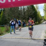 Biegacz przekracza linię mety pod różowo-niebieskim łukiem START, otoczony drzewami i kibicami, czasomierz pokazuje 25 minut.