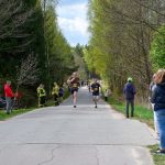 Zawodnicy biegną po asfaltowej drodze przez las otoczony drzewami. Kibice i funkcjonariusze stoją na poboczu. Niebo jest częściowo zachmurzone.