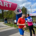 Biegacze przekraczają linię mety pod różowym łukiem startowym. Mężczyzna w czerwonej koszulce sprawdza czas. W tle namiot pomiaru czasu.