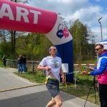 Zawody biegowe na świeżym powietrzu; zawodnik przekracza linię startu-mety, różowy łuk startowy. Tło: drzewa, niebo częściowo pochmurne. Sędzia w niebiesko-różowej kurtce prowadzi zapis wyników.