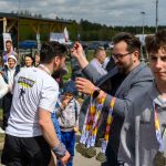 Zdjęcie przedstawia uczestników wydarzenia sportowego. Mężczyzna w okularach rozdaje medale ludziom w tłumie. Tło to zieleń i drewniany budynek.