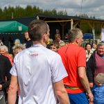 Grupa ludzi na świeżym powietrzu, słońce, zielone tło; centralnie kobieta w czarnej koszulce z białym nadrukiem, dwóch mężczyzn w sportowych koszulkach.