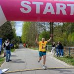 Biegacz w żółtej koszulce przekracza linię startu na szarej drodze. W tle zielone drzewa i niebo. Po bokach obserwatorzy i fotograf.