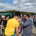 Na zdjęciu mężczyzna w szarej marynarce zakłada medal osobie w żółtej koszulce. Tło to zielone drzewa i niebieskie niebo z chmurami. Wokół widać tłum ludzi, głównie ubranych casualowo.