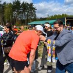 Mężczyzna w szarej marynarce wręcza medale biegaczowi w pomarańczowej koszulce. W tle grupa ludzi i niebo z chmurami. Zieleń i namiot na dalszym planie.