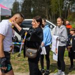 Na zdjęciu kobieta wiesza medal na szyi mężczyzny. W tle namiot i grupa osób na zewnątrz, dzień jest słoneczny. Dominują kolory bieli, niebieskiego i zieleni.