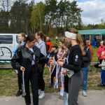 Grupa ludzi, większość młodych kobiet, stoi na zewnątrz, trzymając medale. W tle zielone drzewa i niebo z chmurami. Ludzie są uśmiechnięci i rozmawiają.