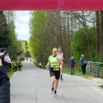 Na zdjęciu biegnący uczestnicy maratonu na drodze otoczonej drzewami. Kobieta w zielonej koszulce uśmiecha się, mijając linię mety. W tle widoczni kibice i zieleń.