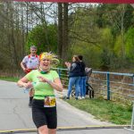 Na zdjęciu kobieta w limonkowej koszulce biegnie przez metę. W tle są drzewa i kibice. Czas na zegarze to 27:22.