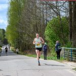 Na zdjęciu biegacz w zielonej koszulce biegnie po asfaltowej drodze w lesie. Po bokach trasy stoją widzowie. W tle widać drzewa i zachmurzone niebo.