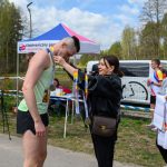 Kobieta zakłada medal biegaczowi w zielonej koszulce. W tle namiot, banery i drzewa. Dzień słoneczny, uczestnicy uśmiechnięci.