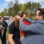 Osoba wręcza medale uczestnikom na zewnątrz. Kolorowe wstążki, ludzie w tle, zielone drzewa. Słoneczny dzień.