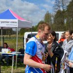 Mężczyzna w stroju biegacza stoi na zawodach. W tle biało-różowo-niebieski namiot i drzewa. Kilka osób obserwuje wydarzenie.