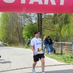 Biegacz przekracza linię mety pod różowym bannerem startowym. Tło z zielonymi drzewami i kilkoma widzami. Zegar pokazuje czas 28:07.