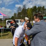 Mężczyzna wręcza medale zawodnikowi na zawodach na tle namiotu. Dookoła stoją widzowie. Niebo jest częściowo zachmurzone.