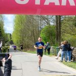 Biegacz w niebieskiej koszulce przekracza linię startu na wiejskiej drodze. W tle drzewa i kibice, zdjęcie robione w słoneczny dzień. Na górze różowy baner z napisem START.