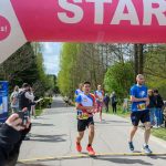 Biegacze startują pod różowo-białym łukiem z napisem START. W tle zielone drzewa i niebo. Obok kibice z aparatami.