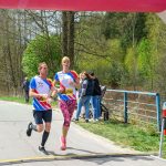 Dwóch biegaczy przekracza linię mety podczas zawodów. Mają na sobie kolorowe stroje sportowe, a tło to zielone drzewa. Obok widać licznik czasu, pokazujący 28 minut 45 sekund.