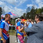 Grupa uczestników biegu w kolorowych strojach, jeden otrzymuje medal. W tle namiot, drzewa i niebo z chmurami.