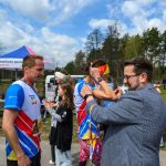 Biegacze w kolorowych koszulkach przy namiocie na tle lasu. Słoneczna pogoda, ludzie wokół robią zdjęcia. Rozdawanie medali.