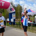 Biegacze przy mecie pod różowym łukiem, z czasem 28:55. Jeden z nich ubrany w biało-niebieski strój, drugi w czarno-biały, obok namiot.
