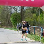 Biegaczka w czarnym stroju przekracza linię mety przy czasomierzu 29:12. W tle zielone drzewa i niebieskie barierki. Baner różowy z białymi literami.