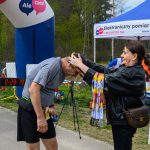 Na zdjęciu kobieta zakłada medal mężczyźnie na mecie biegu. Tło to zielone drzewa i namiot. Widoczna tablica z czasem 29:19.