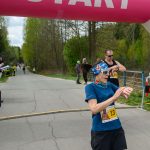 Zawodniczka w niebieskiej odzieży z opaską mija linię mety z czasem 29:20. Nad nią różowa brama startowa. W tle zielone drzewa i widzowie robiący zdjęcia.