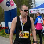 Zawodnik w biegu, numer 110, ubrany na czarno, z medalem na szyi. Tło to zielone drzewa i niebieski dmuchany łuk z napisem. Po lewej licznik czasu, z prawej biały namiot z reklamą.