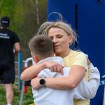 Osoba w żółtej koszulce i chłopiec w bieli przytulają się. W tle są drzewa i niebieska konstrukcja. Obok przechodzi mężczyzna w czerni.