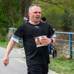 Mężczyzna biegnie w maratonie, ubrany na czarno z białymi akcentami, trzymając numer startowy. Tło z zielonymi drzewami i niebieską barierką. Zdjęcie zrobione na zewnątrz, podczas cieplejszego dnia.