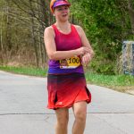 Biegaczka w czerwono-fioletowym stroju sprawdza czas na zegarku. Na żwirowej ścieżce, otoczona drzewami. Nosi żółte buty.