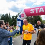 Na zdjęciu mężczyźni kończą bieg pod różowym łukiem startu i mety. Pierwszy z nich ma pomarańczową koszulkę, obok są kibice. W tle widać drzewa i niebieskie niebo.