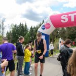 Biegacz przekracza różowy balonowy łuk startowy. W tle zielone drzewa i niebieskie niebo. Wokół stoją kibice.