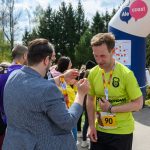 Mężczyzna w żółtej koszulce z medalem, otoczony ludźmi. W tle zielone drzewa i niebiesko-różowy balon. Jasne, słoneczne niebo.