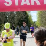 Biegacze na trasie pod różowym banerem START. Dzień słoneczny, drzewa w tle, kibice po bokach dopingują.