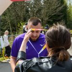 Mężczyzna w fioletowej koszulce na mecie biegu. Kobieta zakłada mu medal. Tło: drzewa, wiosenny dzień.