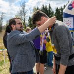 Mężczyzna zakłada medal na szyję uczestnika biegu. Tło stanowi zielone pole i drzewa pod niebieskim niebem. Wokół widać innych uczestników.