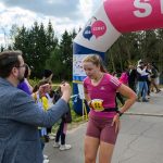 Kobieta w różowym stroju biegnie na zawodach. Mężczyzna wręcza jej medal za linią mety. W tle drzewa i ludzie.